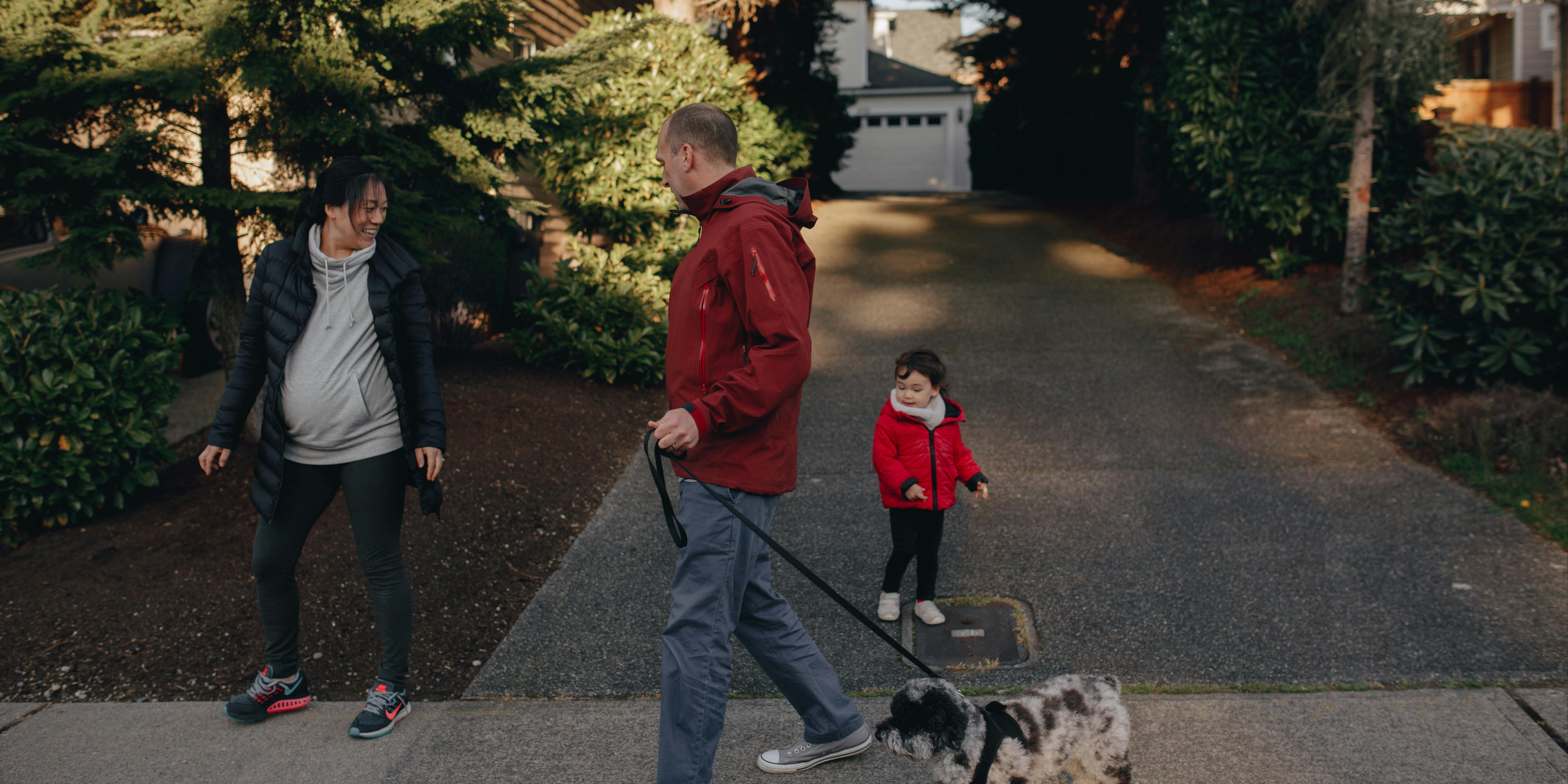 a couple walks a small dog with their toddler in a driveway